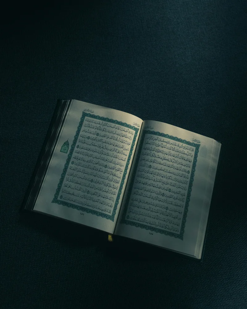 An open Qur'an showing two pages of Arabic text, softly lit on dark cloth
